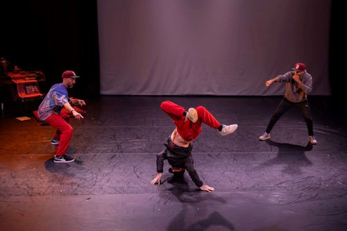 A dancer is captured as he breakdances centre stage. Bobak and another performer stand to either side of the dancer. 