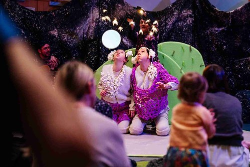 Two performers dressed as sheep gaze upward in wonder, surrounded by children and parents watching attentively in a cosy, immersive theatre space. A performer in the background holds a glowing umbrella-like prop overhead.
