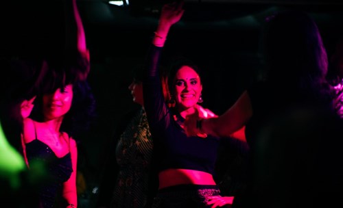 A group of women dancing joyfully in a dimly lit space with vibrant pink and purple lighting. The central woman, wearing a black crop top and traditional jewellery, smiles brightly with one arm raised in celebration