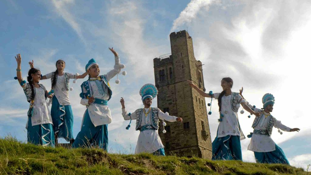 Virsa Bhangra dancers in blue and white traditional costumes perform outdoors on Castle Hill, with Victoria Tower rising behind them beneath a bright blue sky.