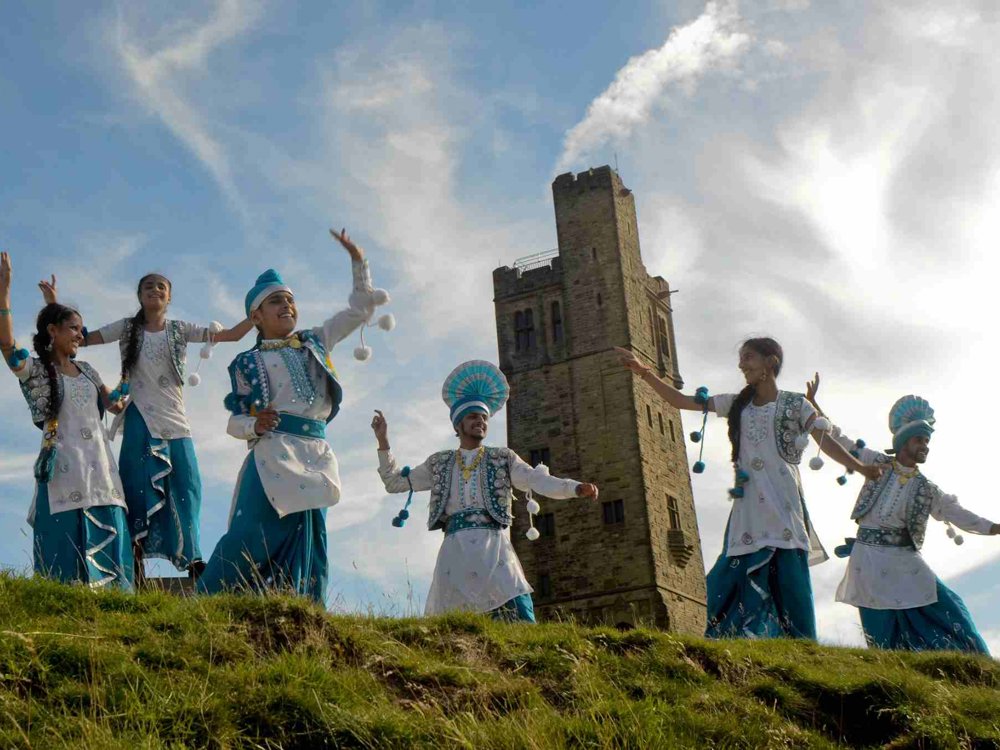 Virsa Bhangra dancers in blue and white traditional costumes perform outdoors on Castle Hill, with Victoria Tower rising behind them beneath a bright blue sky.