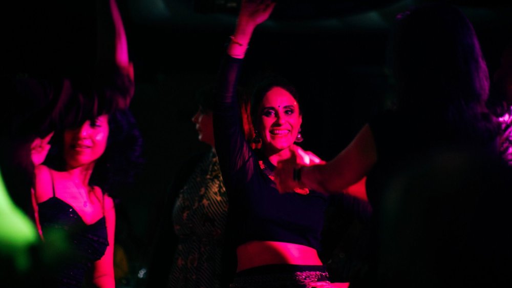 A group of people dance together in a dimly lit venue, illuminated by vibrant pink and purple club lights. One smiling person in the centre raises an arm in the air while others dance around them, creating a lively and energetic party atmosphere.