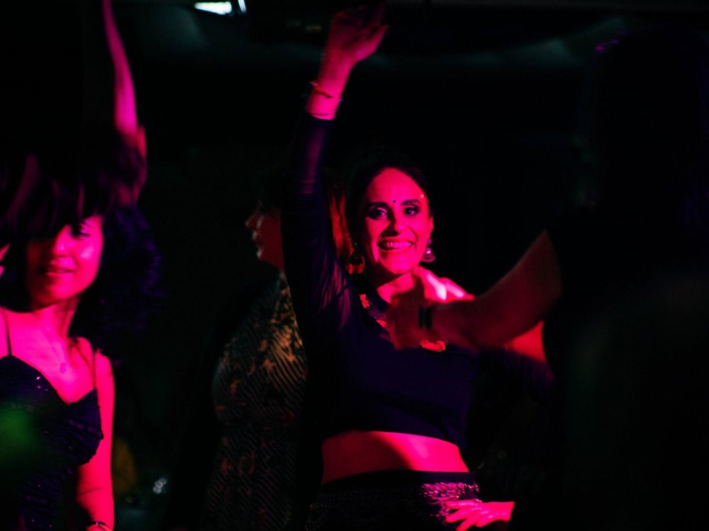 A group of people dance together in a dimly lit venue, illuminated by vibrant pink and purple club lights. One smiling person in the centre raises an arm in the air while others dance around them, creating a lively and energetic party atmosphere.