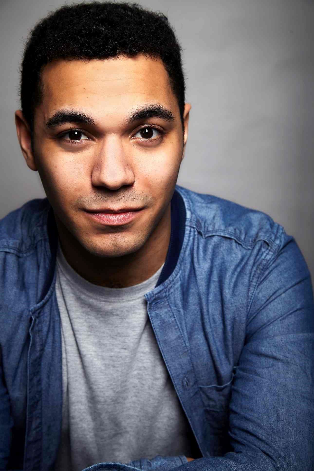 Reece Carter: Man with short curly hair wearing a light grey T-shirt and blue jacket, smiling slightly at the camera.