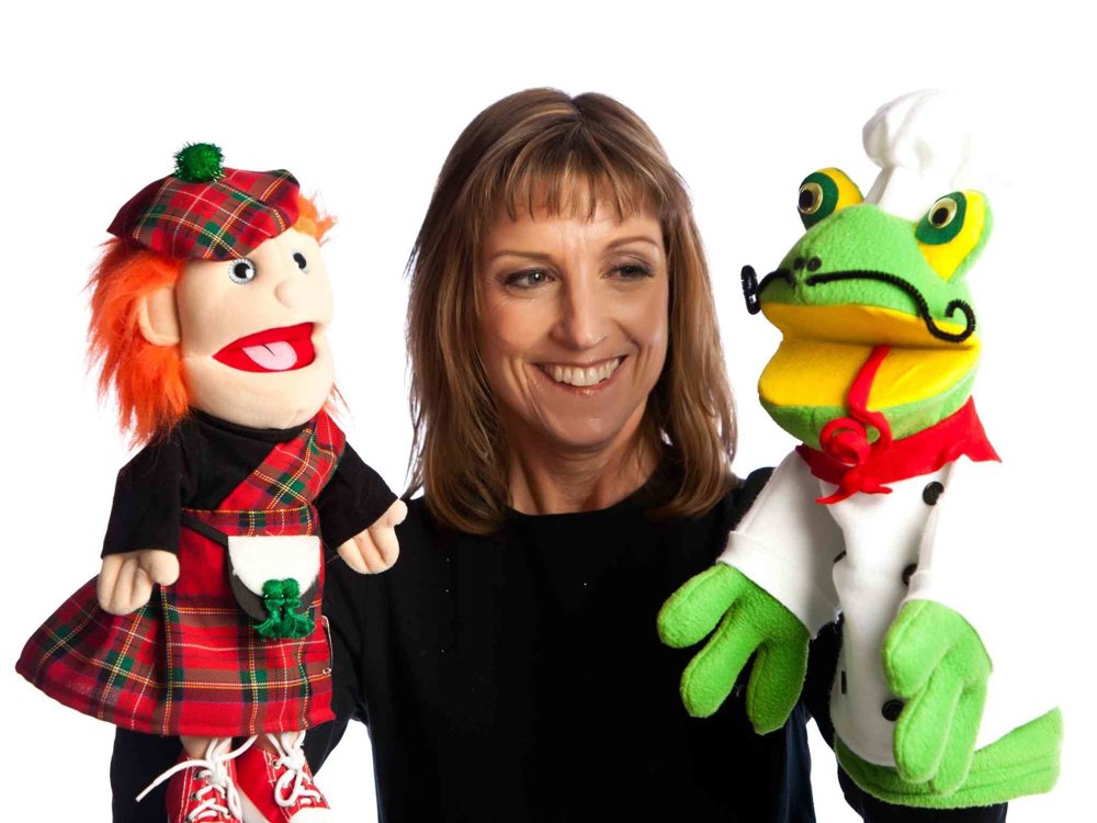 A smiling woman dressed in black holds two colourful hand puppets against a plain white background. One puppet wears a red tartan Scottish outfit with red hair and boots, while the other is a bright green frog dressed as a chef with a white hat and curled moustache.