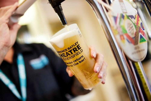 A bartender pours a pint of beer from a tap into a reusable plastic cup branded with "Lawrence Batley Theatre Huddersfield." The bartender wears a black uniform with a lanyard, and the background is blurred, showing a busy bar setting.