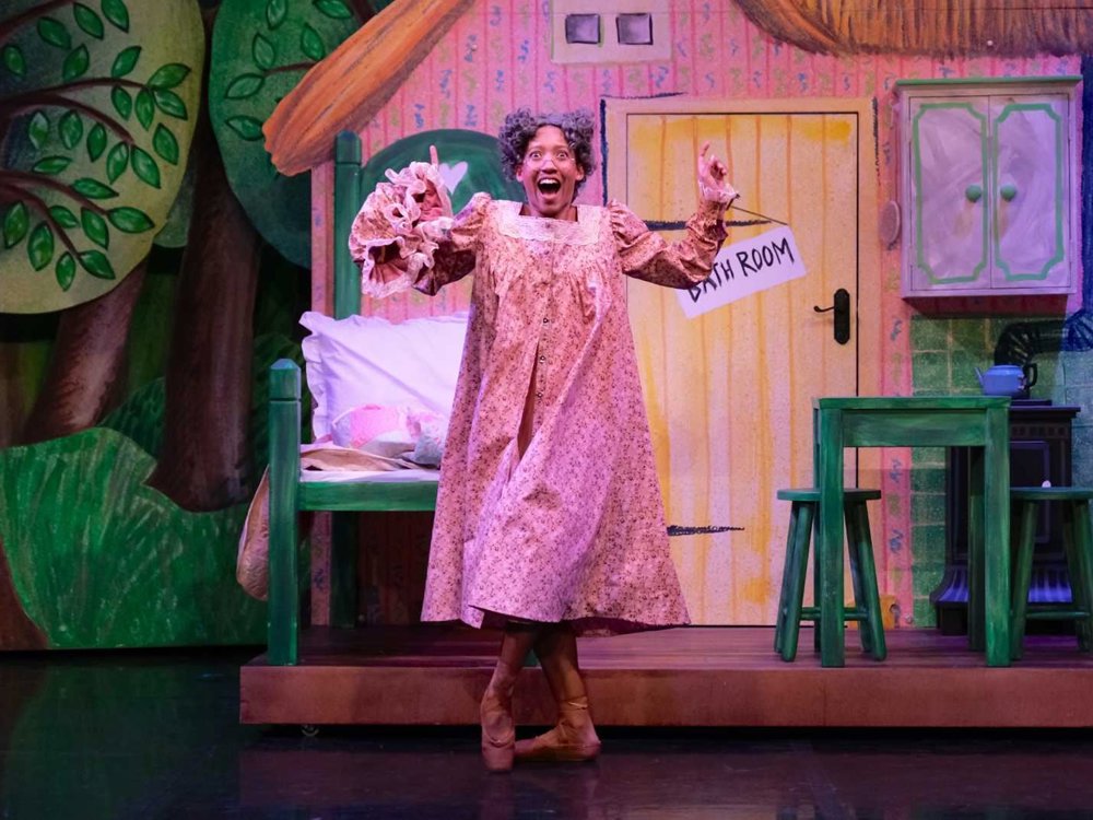 A performer portraying Grandma stands on pointe beside a bed and table in a cottage set, wearing a floral nightgown and smiling joyfully.