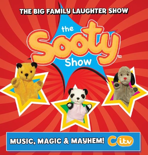 Promotional image for The Sooty Show with a bright red starburst background. At the top, bold text reads “The Big Family Laughter Show.” In the centre, the logo says “The Sooty Show” with “Sooty” in large yellow lettering. Three puppet characters appear in yellow star frames: Sooty (a yellow bear), Sweep (a grey dog with black ears), and Soo (a panda in a pink dress).