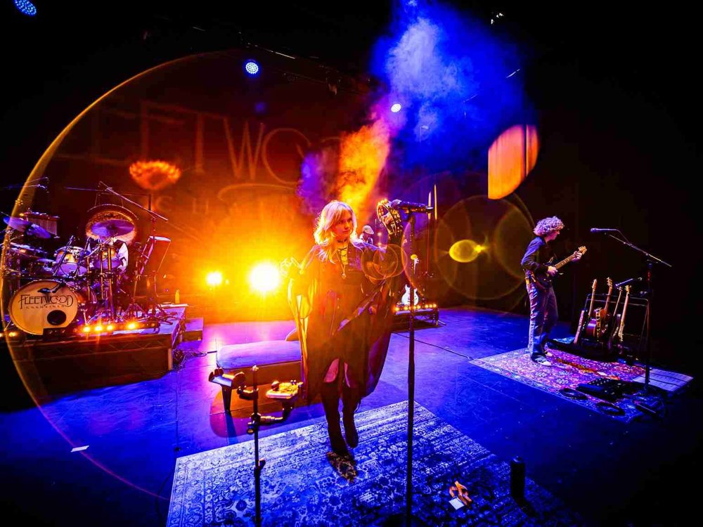 Live performance of Fleetwood Unchained with the drummer on the left, a female vocalist centre stage under warm orange lighting, and a guitarist playing on the right. Bright stage lights and lens flares fill the background.