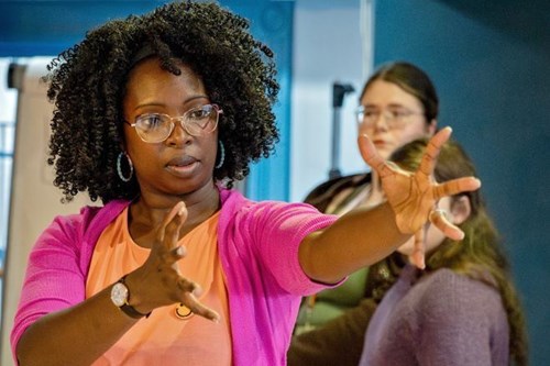 A woman with glasses, wearing a pink cardigan and peach shirt, gestures expressively with outstretched hands during a rehearsal.