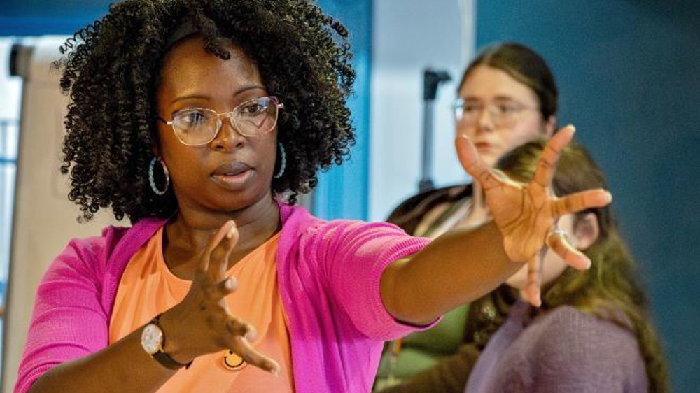 A woman with glasses, wearing a pink cardigan and peach shirt, gestures expressively with outstretched hands during a rehearsal. 