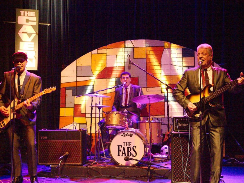 A three-piece band performing on stage in suits, with one playing guitar, one on drums, and one on bass. A drum kit with “The Fabs” logo is centre stage, and behind them is a colourful mural backdrop with the words “The Cavern” lit up to the side.
