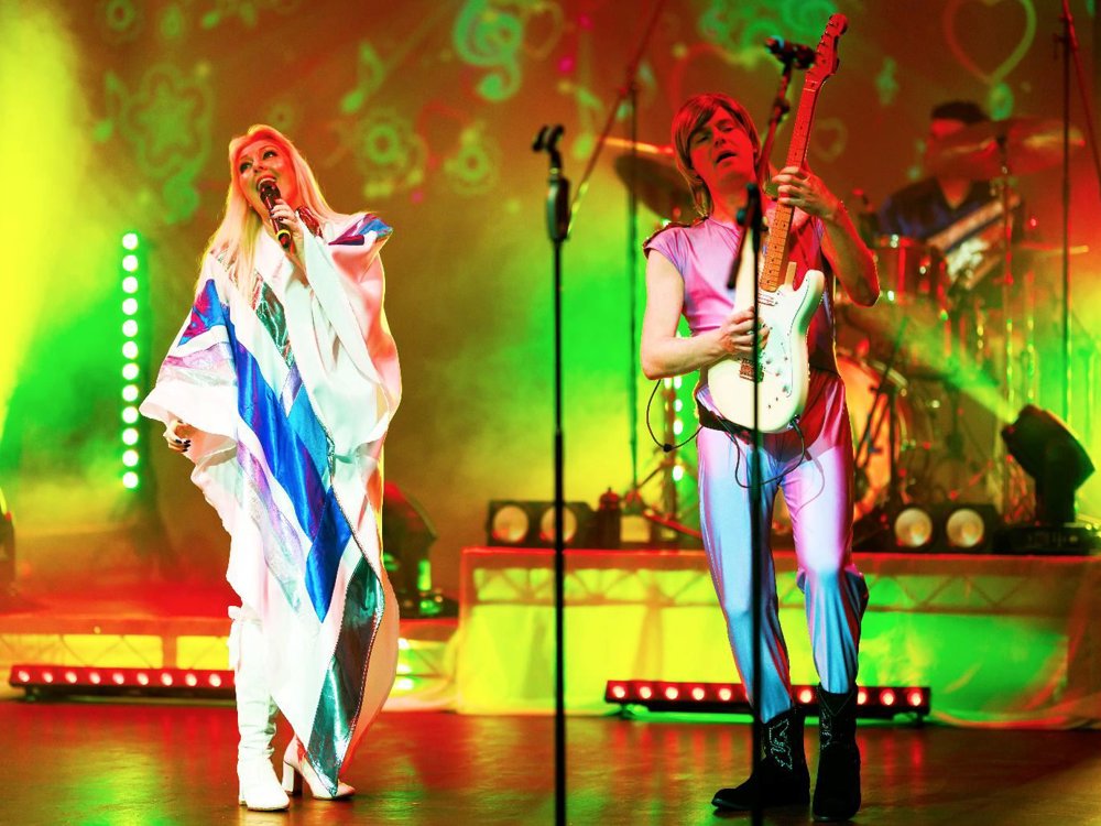 A singer and guitarist perform together under vivid green and yellow stage lights, with the drummer playing behind them during an energetic concert number.