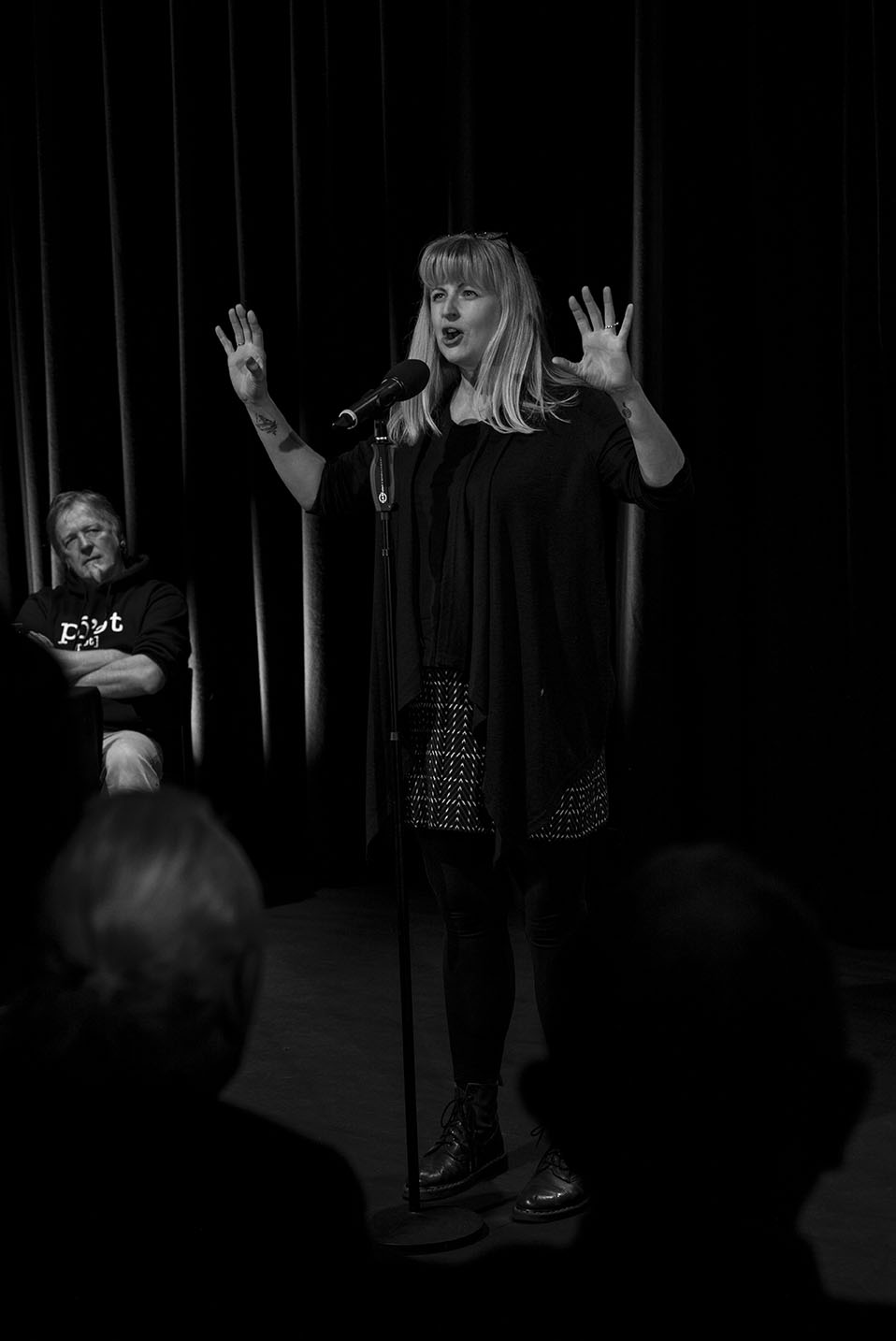 A black-and-white photograph of a woman performing spoken word poetry at a microphone on a theatre stage. She gestures expressively with both hands raised while addressing the audience. A seated judge or host watches from the side of the stage, and audience silhouettes fill the foreground, creating an intimate live performance atmosphere.
