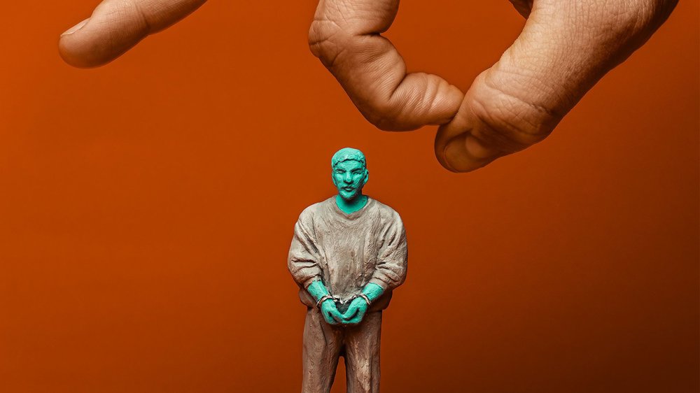 A small clay figurine of a handcuffed person stands on an orange surface, with a large human hand reaching down from above as if about to knock it over. 