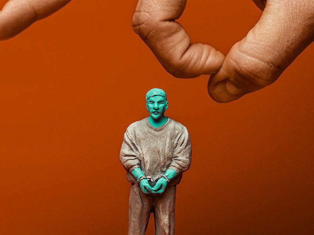 A small clay figurine of a handcuffed person stands on an orange surface, with a large human hand reaching down from above as if about to knock it over. 