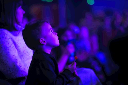 A young boy sits in the audience at Lawrence Batley Theatre, gazing up in awe at the stage, illuminated by purple and blue lights, while holding a red lollipop in his hand.