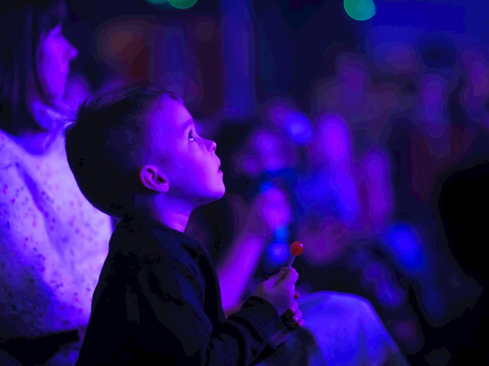 A young boy sits in the audience at Lawrence Batley Theatre, gazing up in awe at the stage, illuminated by purple and blue lights, while holding a red lollipop in his hand.