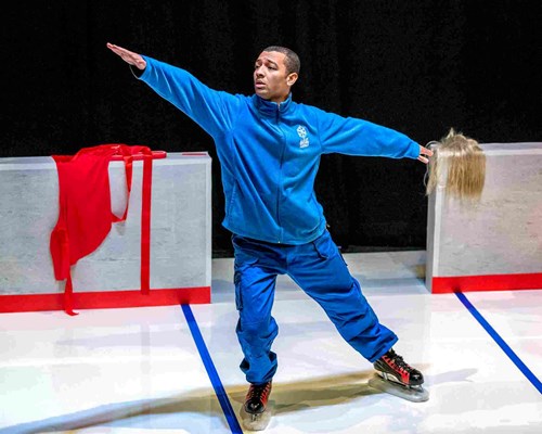 The caretaker (a human!) mimics ice skating wearing a full, blue tracksuit and large boots, like ice skates. He stands on a white floor like that of an ice rink. 