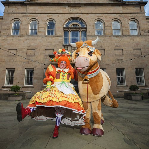 A photograph of a pantomime dame and cow in front of the theatre.