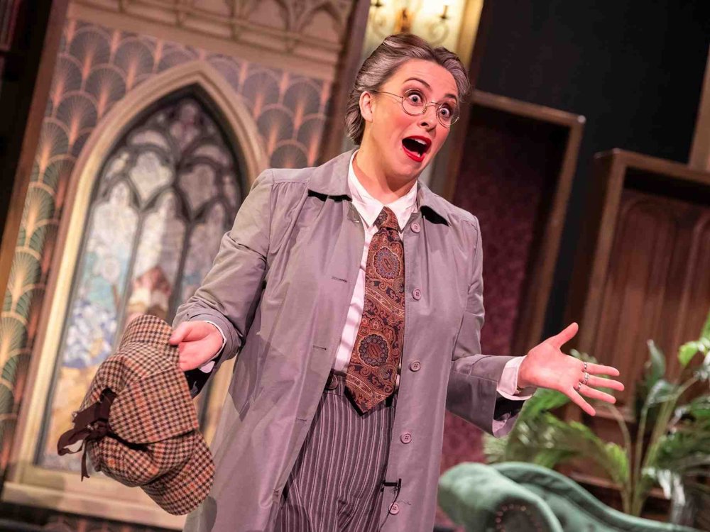 An actor in a grey trench coat and paisley tie performs expressively on stage, holding a deerstalker hat with a shocked or surprised expression