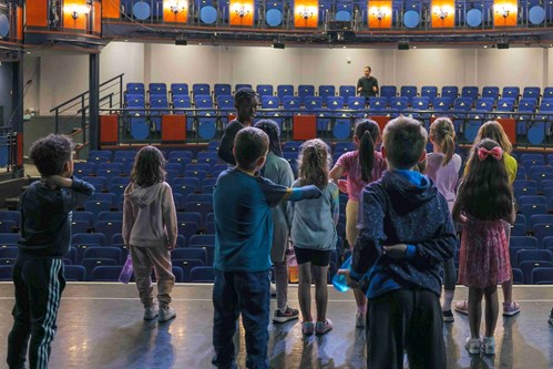 A group of children are standing on the Lawrence Batley Theatre stage with their backs to the camera, looking out towards rows of seats
