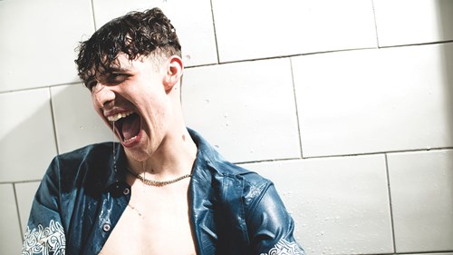 A young man with wet hair is standing against a tiled wall, wearing a blue shirt. He is yelling passionately, with water dripping from his mouth.