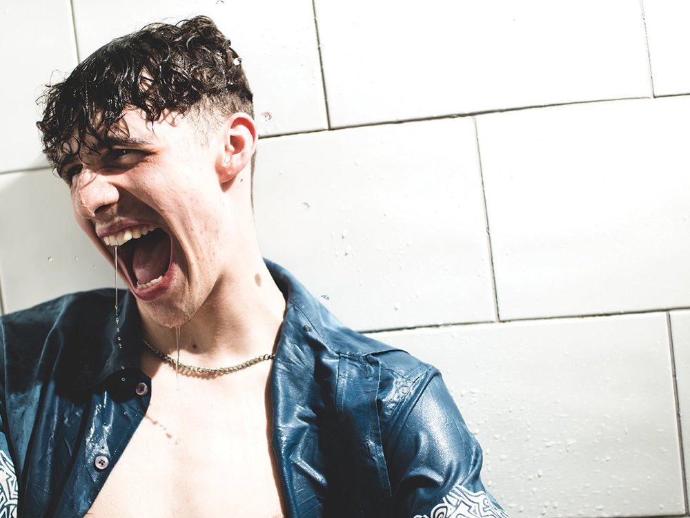 A young man with wet hair is standing against a tiled wall, wearing a blue shirt. He is yelling passionately, with water dripping from his mouth.