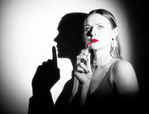 A black and white image of a women applying bright red lipstick, the only colour on the image. She looks glamourous with jewelled earrings and a necklace to match. Behind her, is a silhouette of a man with a finger to his lips, shushing. 