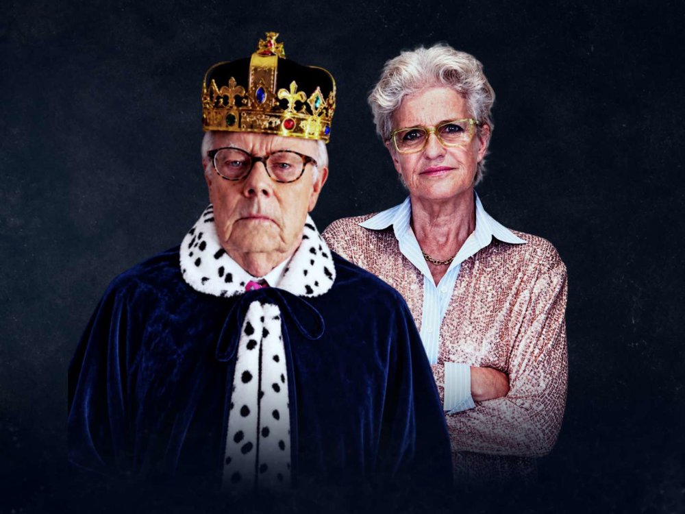 An older man wearing a gold crown and dark blue royal robe with white spotted fur trim stands in front of an older woman with short grey hair, glasses and a pink patterned jacket, against a dark background.