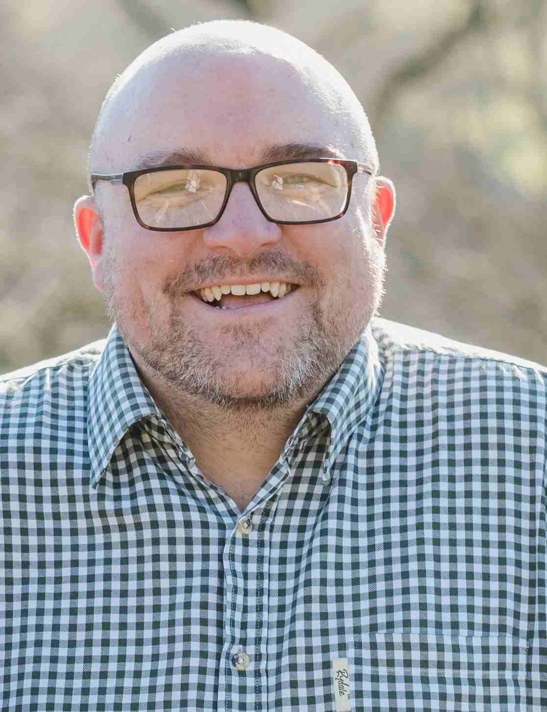 Pete Toon: Smiling man with glasses and a shaved head, wearing a green and white checkered shirt, photographed outdoors in natural light.