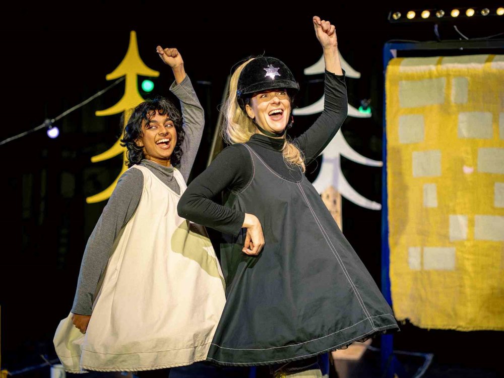 Two performers dance joyfully with raised arms on a brightly lit stage. One wears a toy police-style helmet and blonde ponytail, adding a playful twist to the fairytale world.