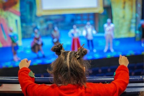 A young child with styled pigtails and wearing a red jumper leans over a theatre balcony, watching a group of costumed performers on stage during a brightly lit pantomime show.