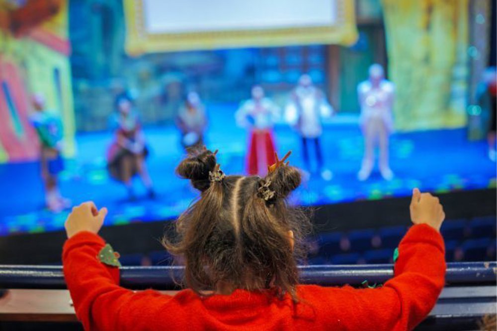A young child with styled pigtails and wearing a red jumper leans over a theatre balcony, watching a group of costumed performers on stage during a brightly lit pantomime show.