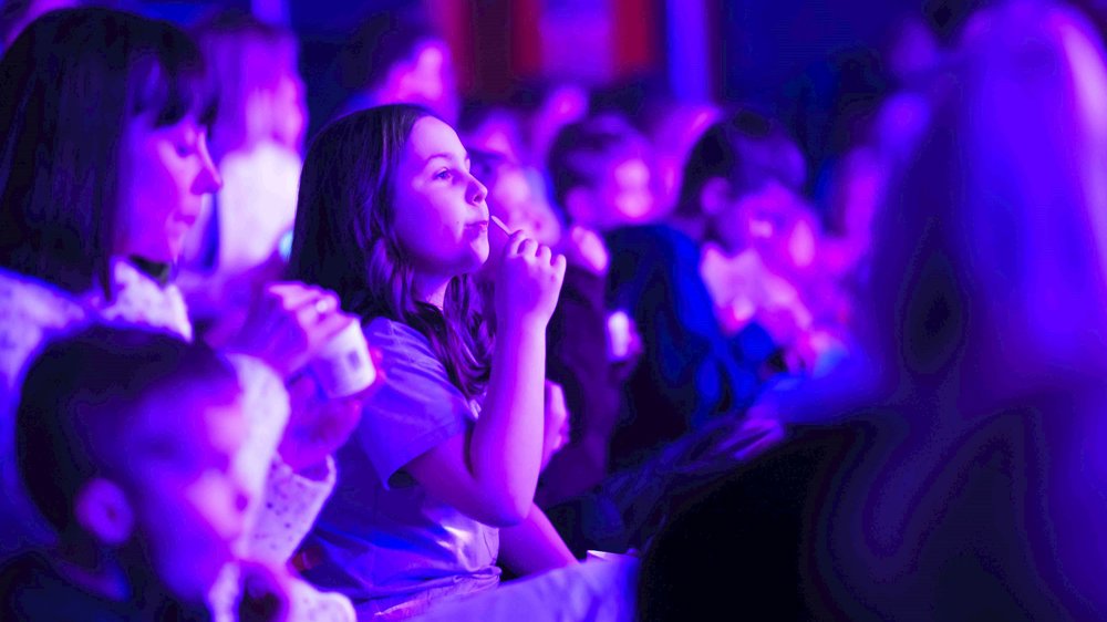 A captivated audience of children and adults sits bathed in vibrant purple lighting, watching a live performance.