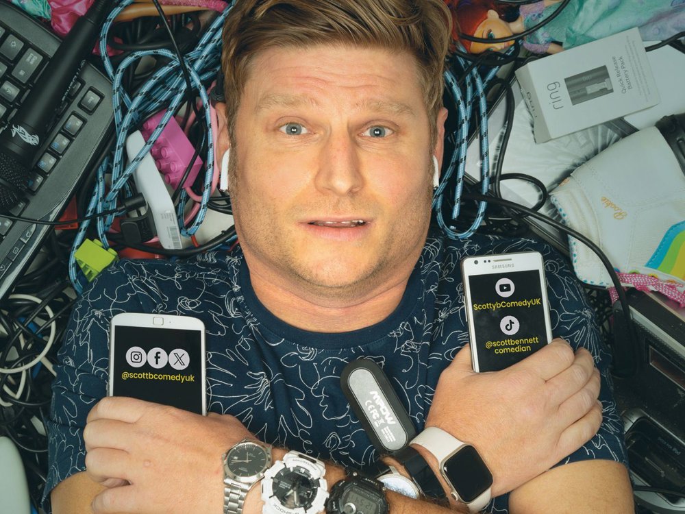 Comedian Scott Bennett lies on his back surrounded by a chaotic tangle of tech gadgets, wires, and miscellaneous items including a microphone, keyboard, smartwatches, and a roller skate. He holds two smartphones displaying his social media handles: @scottbcomedyuk on Instagram, Facebook, and X, and @scottbennettcomedian on TikTok and ScottybComedyUK on YouTube. His expression is humorous and overwhelmed, with earbuds in and multiple smartwatches on his wrists.