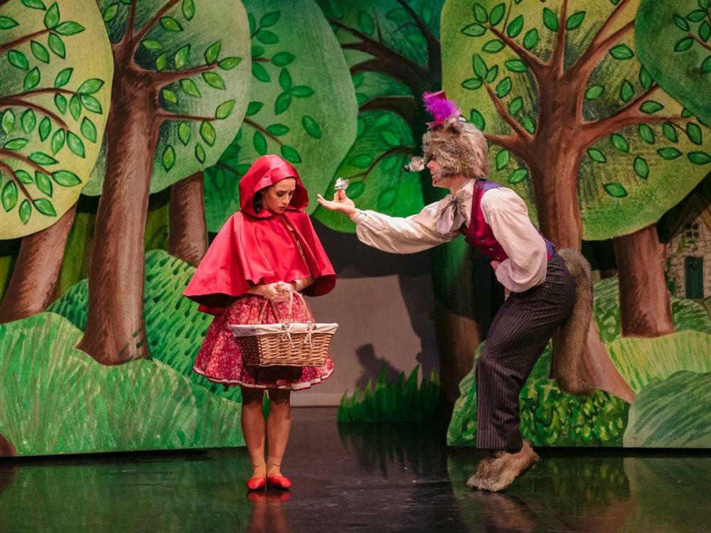 Little Red Riding Hood, wearing a red cape, meets the Wolf in a waistcoat and top hat on stage in front of a colourful woodland set.