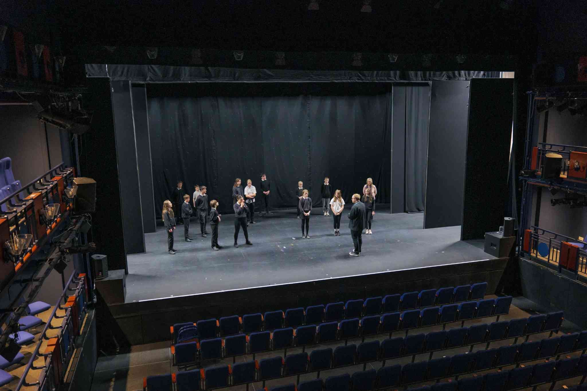 “A group of young people standing on a theatre stage during a rehearsal or workshop, viewed from the auditorium with rows of empty seats in the foreground.”