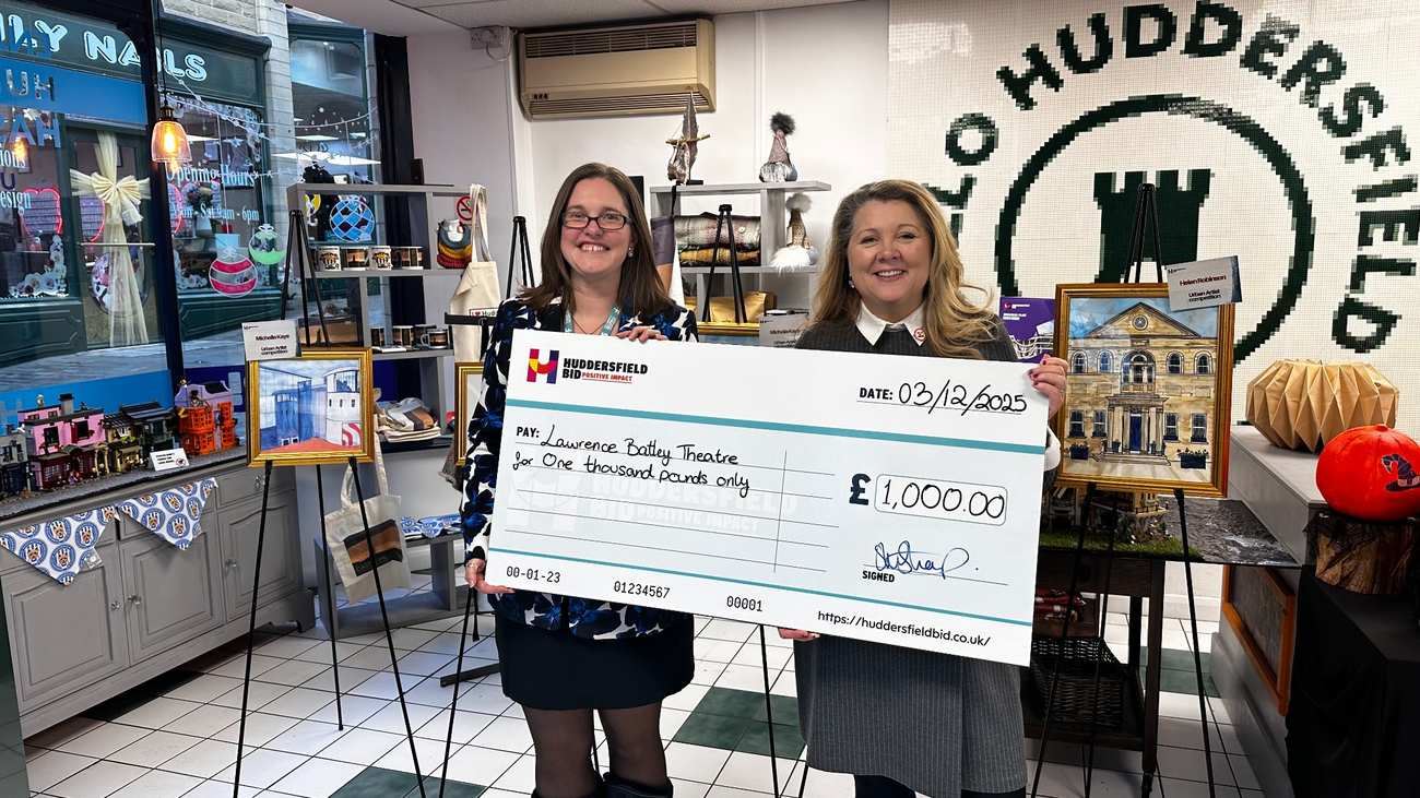 Two women stand inside a shop space holding a large Huddersfield BID cheque for £1,000 made payable to Lawrence Batley Theatre, surrounded by displayed artwork and creative merchandise.