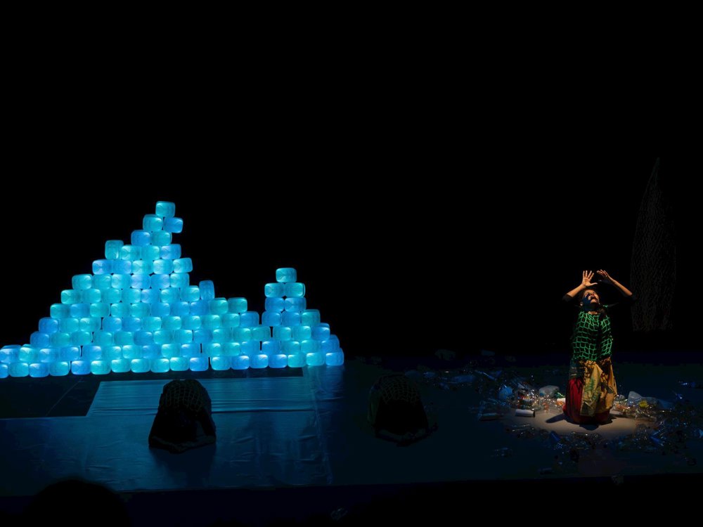 One dancer reaches upward while surrounded by scattered plastic waste on the floor. Two other performers crouch nearby, with illuminated blue plastic-container structures forming the backdrop.