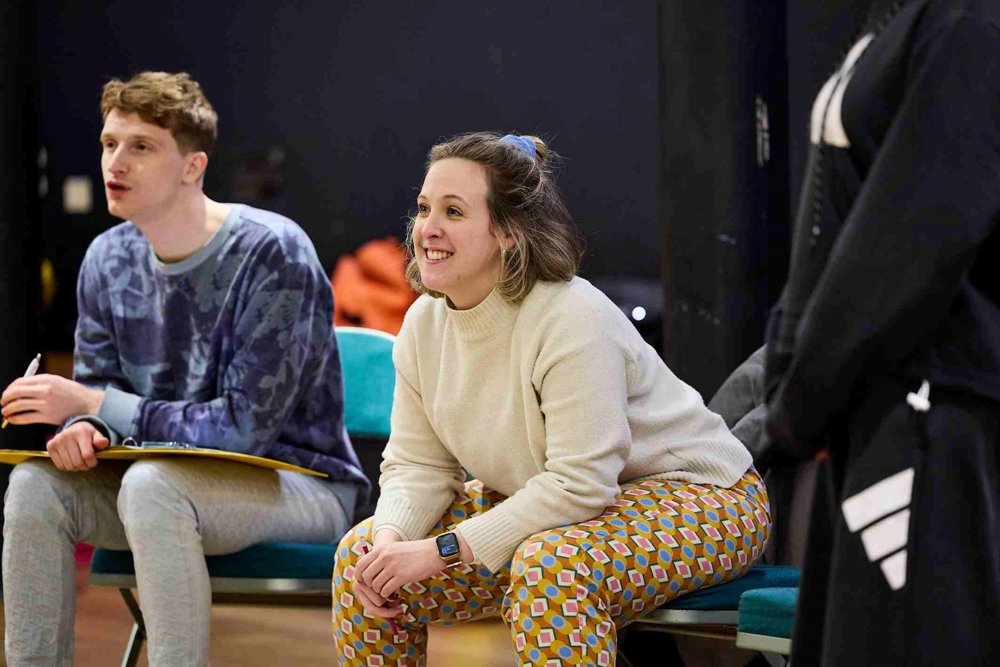 Director Olivia Race seated and smiling during a rehearsal. She is wearing a cream jumper and colourful patterned trousers, with her hair tied back and a smartwatch on her wrist. The background shows a rehearsal space with chairs and props.