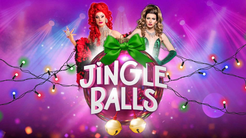 Colourful Christmas-themed promotional artwork for Jingle Balls, featuring two glamorous drag performers, one with bright red hair and one with brunette hair, posed behind a giant pink bauble tied with a green bow. The title “Jingle Balls” appears prominently in large white lettering, surrounded by festive string lights and golden bells against a vibrant pink and purple background.