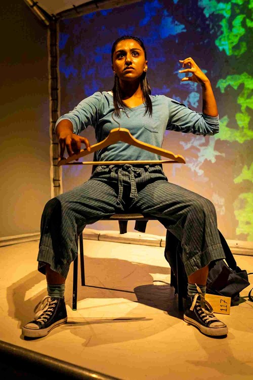 Sitting on a chair, a young person mimics playing a cello using a wooden coat hanger as a bow. They are wearing a blue long sleeved tshirt and blue, striped trousers. 