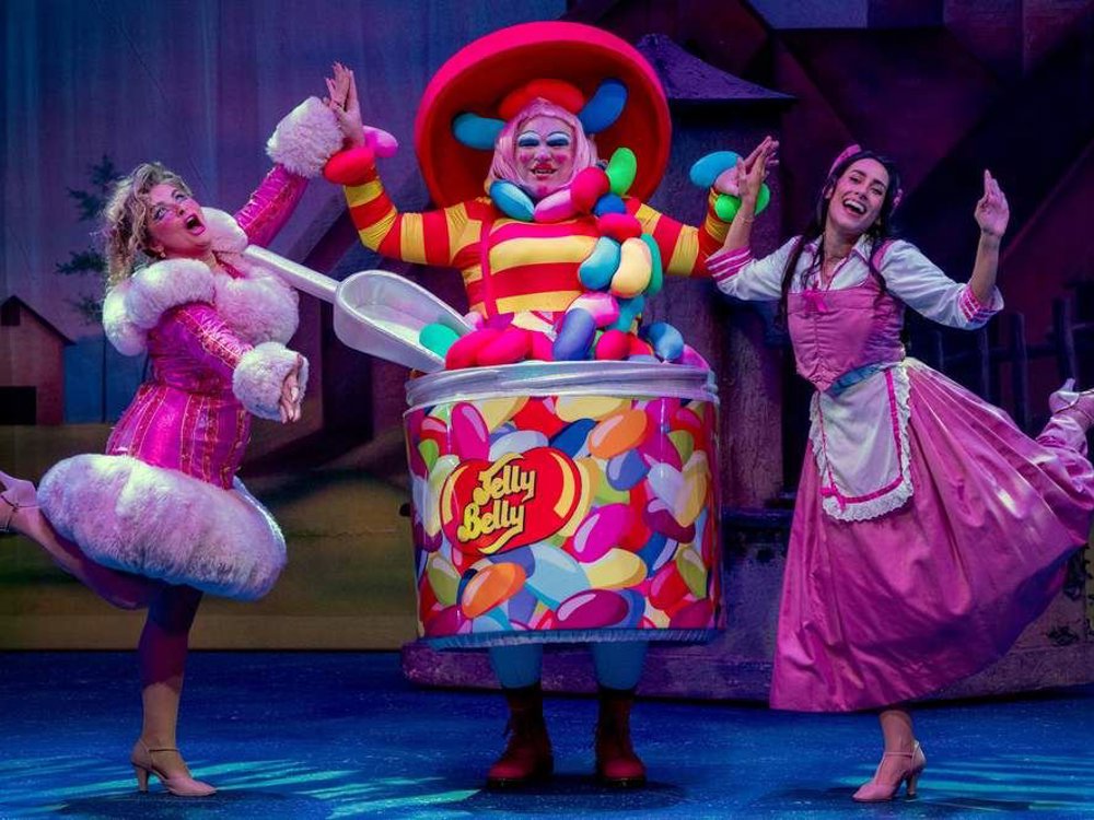 Three pantomime characters pose joyfully on stage, including a performer dressed as a giant Jelly Belly jar, flanked by two performers in pink costumes, all mid-dance against a theatrical backdrop.