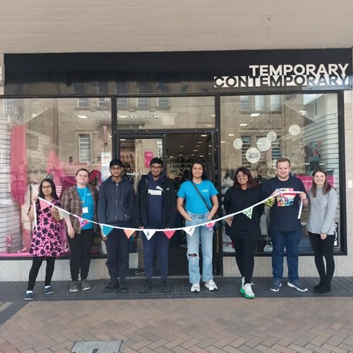 8 young people stand outside a shop holding colourful bunting they have made