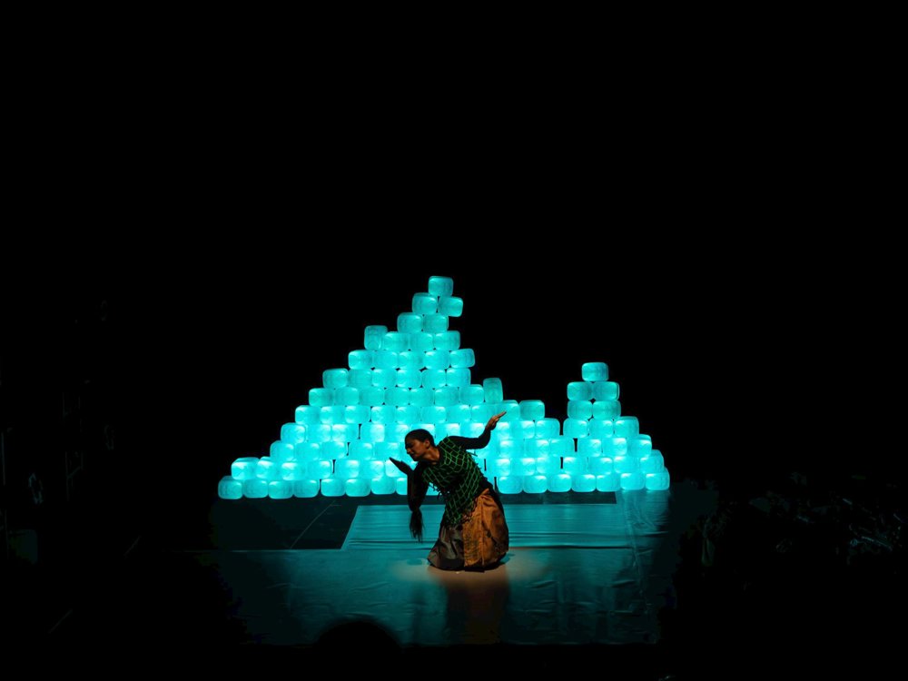 A solo dancer kneels in a spotlight on a dark stage, framed by a glowing wall of stacked plastic containers illuminated in blue-green light.