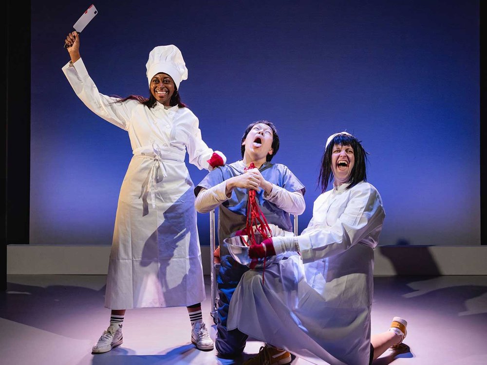 An image of a theatrical scene, a smiling performer dressed as a chef raises a cleaver while two others in medical-style outfits laugh dramatically, one holding a bowl filled with bright red prop intestines.