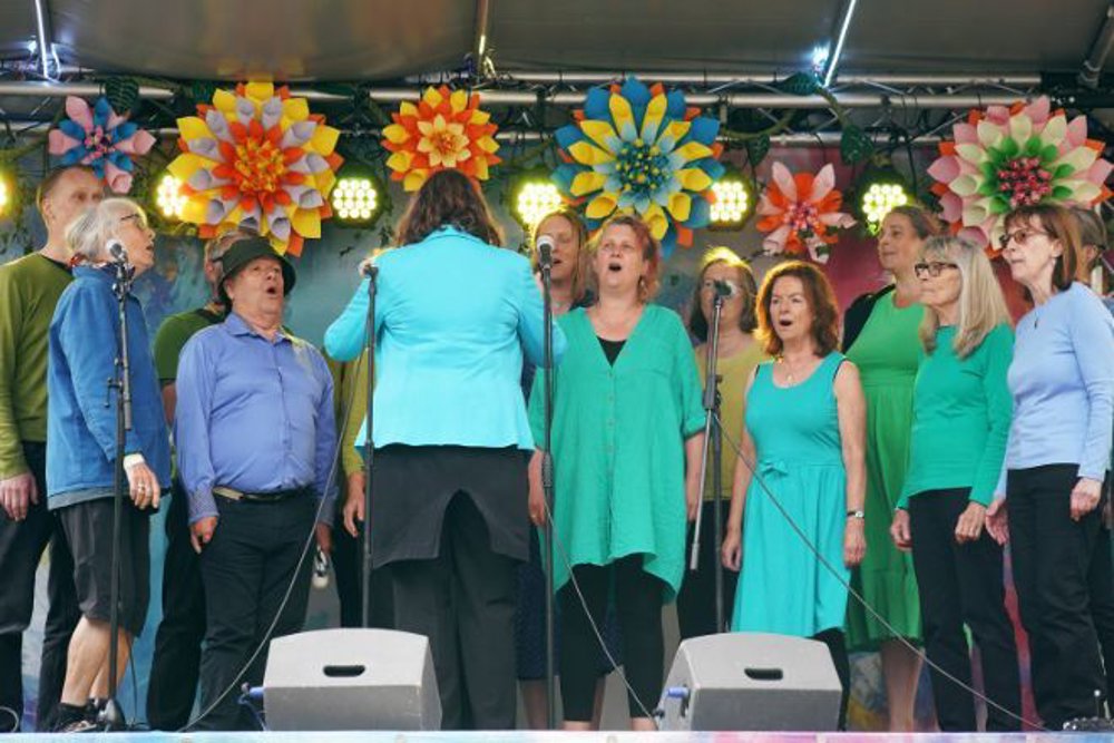 A choir performing on an outdoor stage, led by a conductor wearing a bright blue jacket. The choir members, dressed in shades of green and blue, sing in unison under vibrant floral decorations and stage lighting