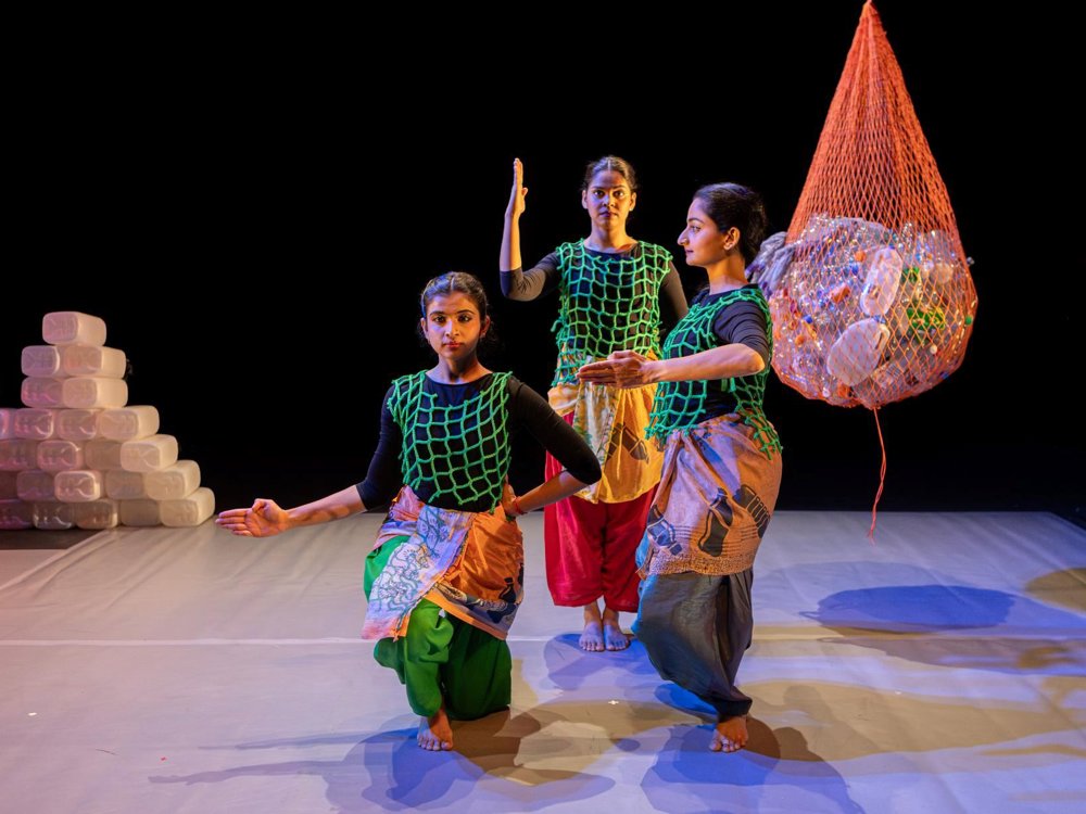 Three dancers hold stylised poses on stage wearing green net-like costumes. A large suspended net filled with plastic bottles hangs beside them, with stacks of plastic containers visible in the background.