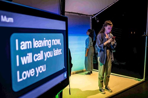 A screen shows a text to Mum. The text reads 'I am leaving now, I will call you later. Love you'. Beside the screen stands a young person looking at their mobile phone. They are wearing jeans and an oversized, blue denim shirt. 